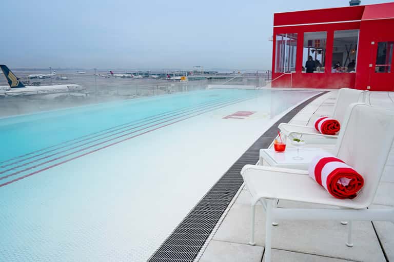 Rooftop lap pool with red cabana building, city airport views, and loungers with red towels