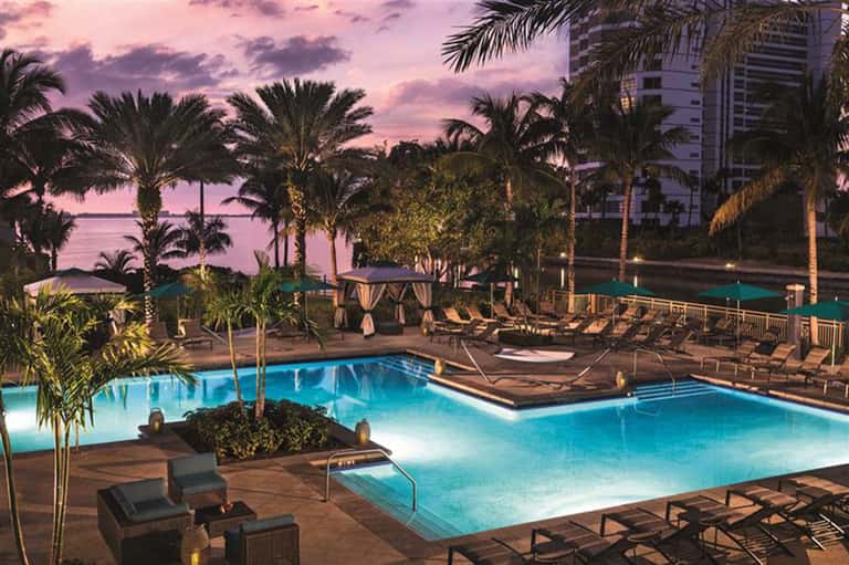 Illuminated resort pool complex at dusk with palm trees, loungers, and waterfront backdrop