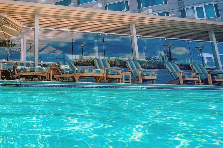 Resort pool with striped loungers, covered pergola, and ocean city view behind glass barrier