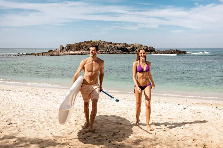 Fit couple on sandy beach holding paddleboards with ocean and rocky island in background