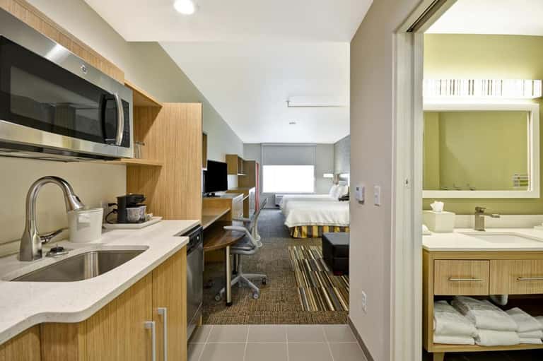 Modern hotel suite showing kitchenette, living area, bedroom with striped bedding, and bright bathroom with yellow accents