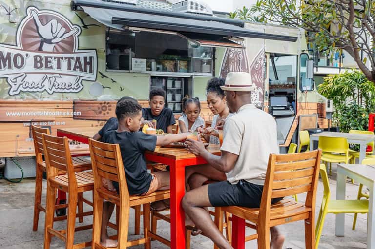Family enjoying food truck meal outdoors at colorful tables near Mo'Bettah food truck
