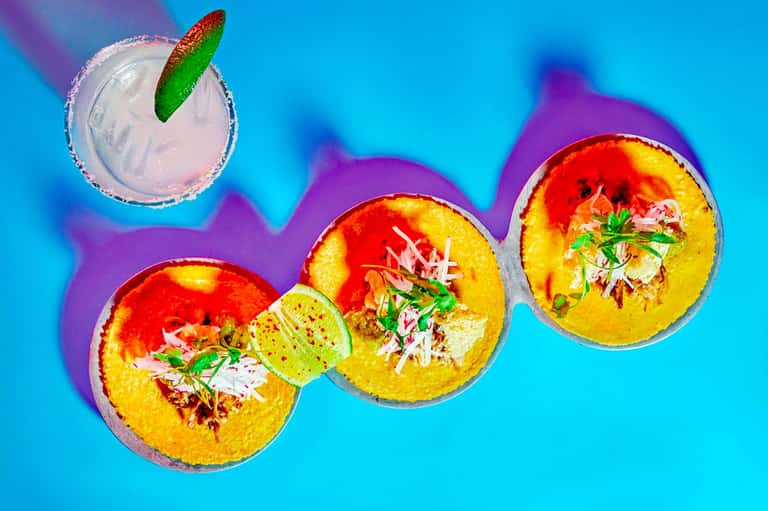 Three colorful tropical bowls with yellow rim, lime garnish, and pink drink on blue background