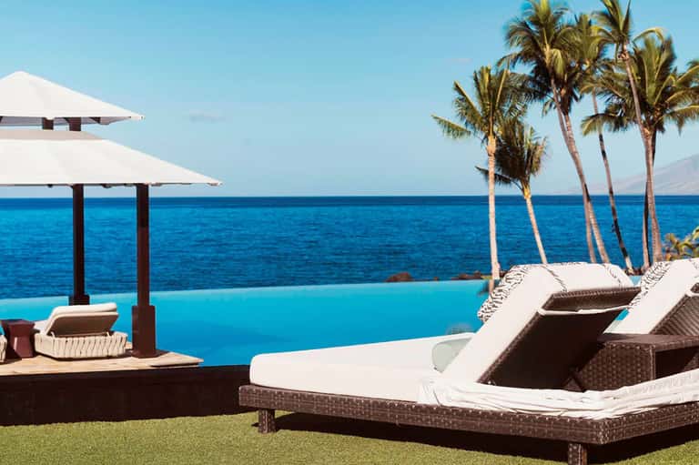 Oceanfront lounge chairs with white umbrellas overlooking infinity pool and palm trees