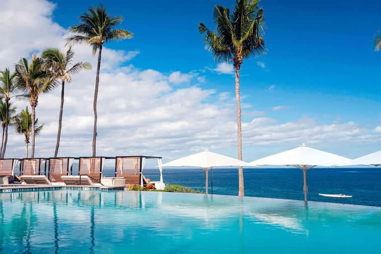 Infinity pool overlooking ocean with palm trees, white umbrellas, and ocean horizon