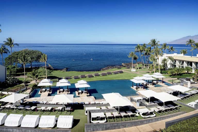 Oceanfront resort pool with white umbrellas, loungers, green lawn, and mountain seascape views