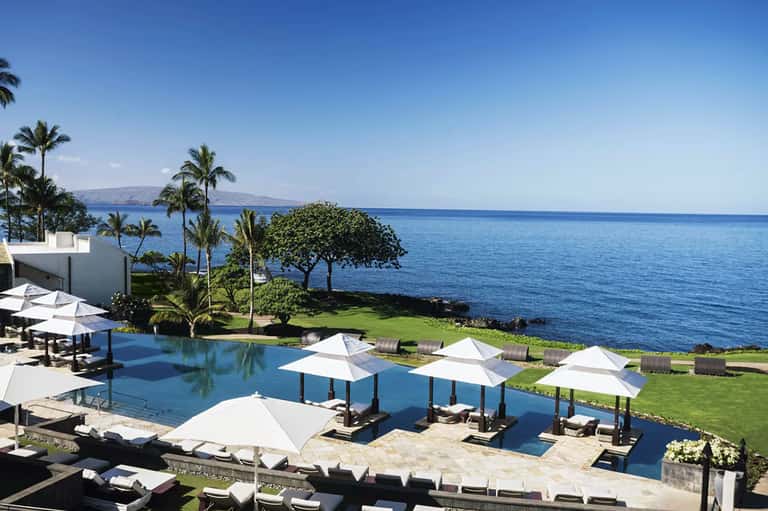 Luxury oceanfront infinity pool with white umbrellas, manicured lawn, and distant island view