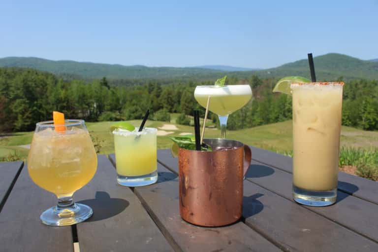 Four colorful cocktails on wooden deck overlooking forested mountain landscape