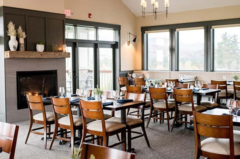 Elegant dining room with fireplace, wood chairs, white tablecloths, and panoramic windows with forest view