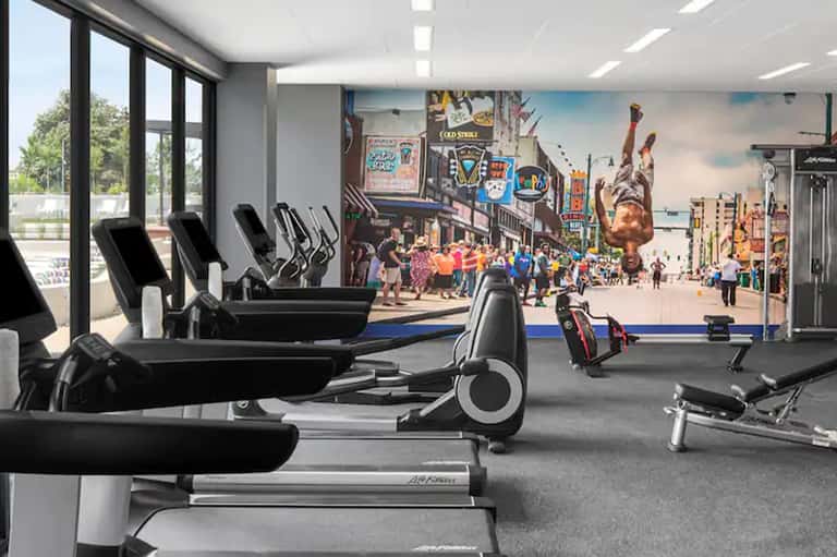 Hotel fitness center with cardio equipment, large windows, and vibrant wall mural of urban scene