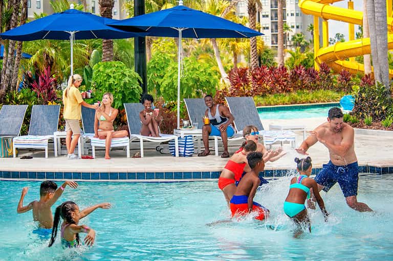 Family-friendly pool area with adults and children playing under blue umbrellas and yellow waterslide