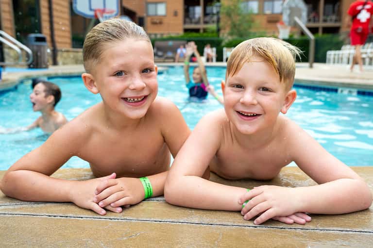 Two young boys smiling at pool edge with water and other swimmers in background