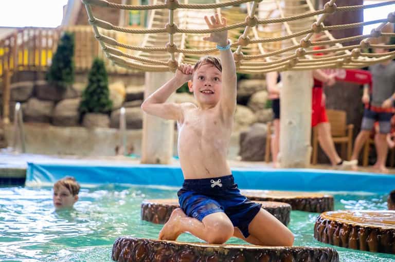 Young boy playing on rope obstacle course over shallow pool water at resort recreation area