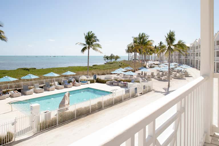 Beachfront resort pool surrounded by lounge chairs, umbrellas, palm trees, and ocean views with white railing