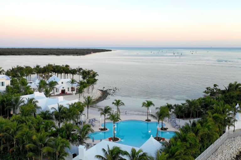 Luxury beachfront resort with white buildings, blue pool, palm trees, and calm ocean waters