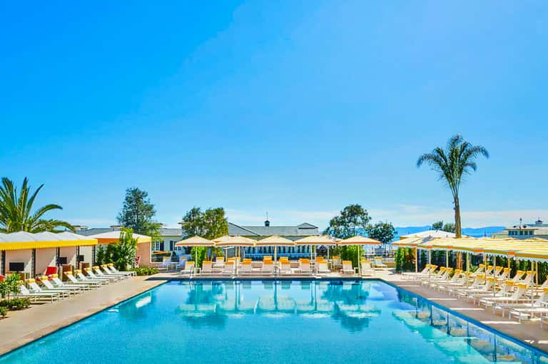 Resort swimming pool with yellow umbrellas, lounge chairs, palm trees, and clear blue sky.