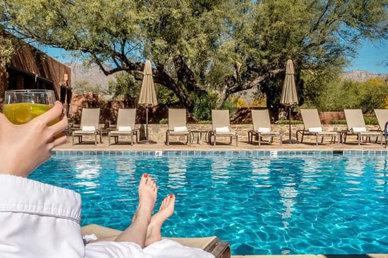 Resort pool lounge with guest holding drink, desert mountains, and shaded seating area