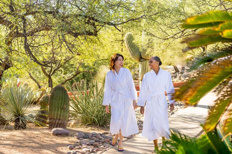 Two women in white robes walking through desert garden with cacti and stone pathways