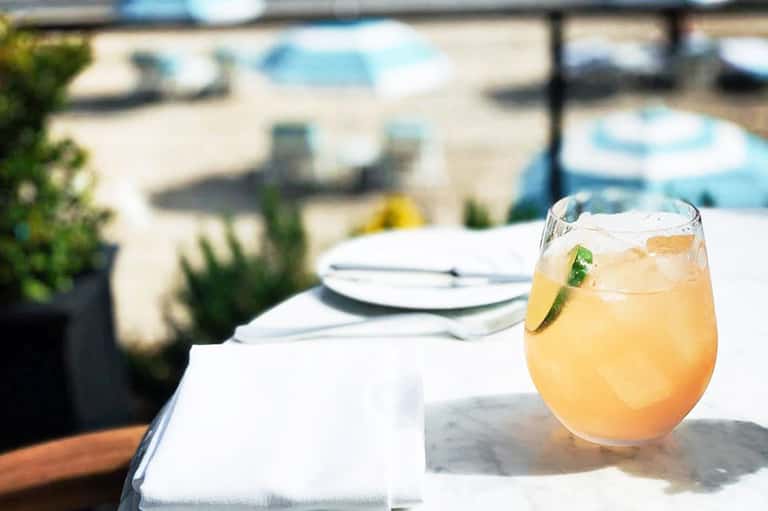 Cocktail with lime garnish on oceanfront dining table with beach umbrellas in background