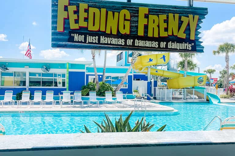 Bright turquoise resort pool with yellow waterslide, lounge chairs, palm trees, and feeding frenzy sign