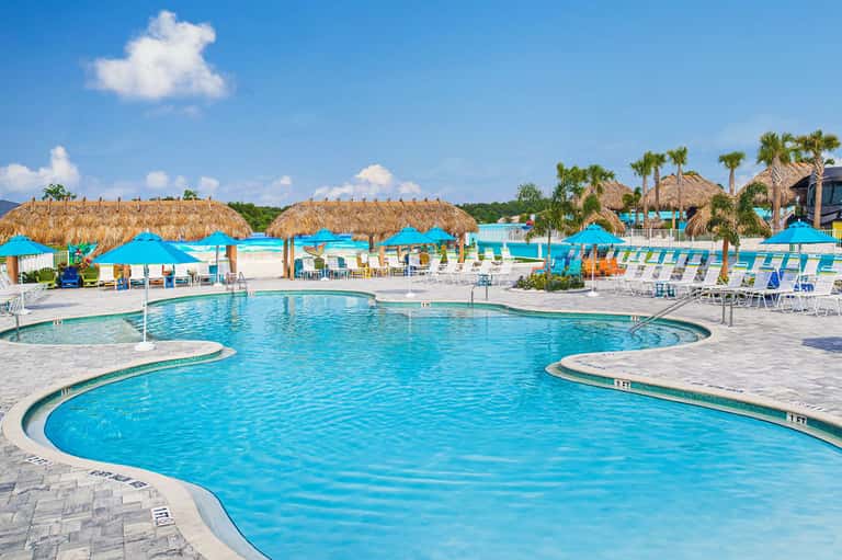 Resort pool with blue cabanas, thatched umbrellas, palm trees, and tropical landscape.