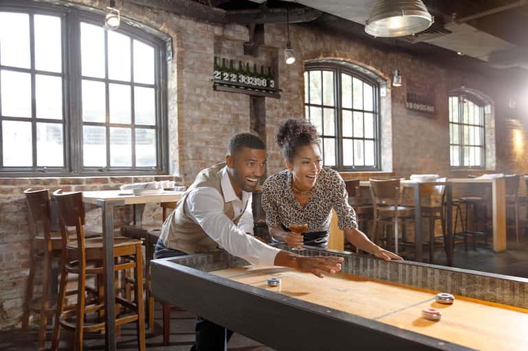 Couple playing shuffleboard in industrial-style bar with exposed brick, large windows, and wooden tables