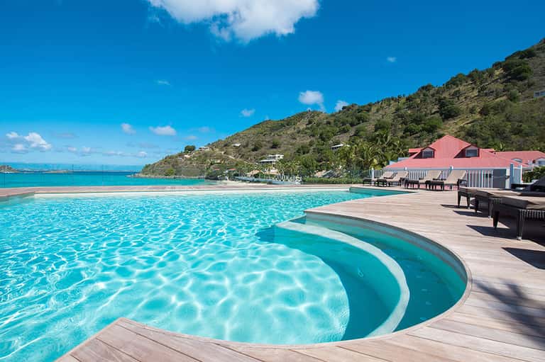 Curved lagoon-style pool with hot tub, red-roofed building, and hillside ocean view backdrop