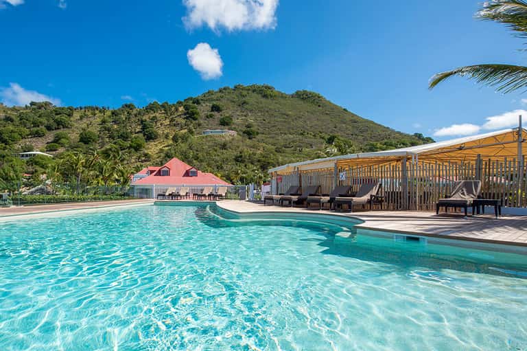 Turquoise pool with wooden loungers under shelter, pink building, and forested hillside backdrop