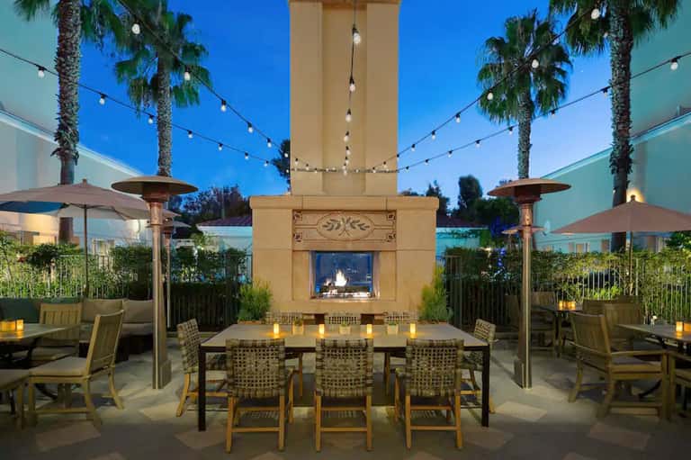 Elegant outdoor dining terrace with fireplace, string lights, palm trees, and formal table setting at dusk