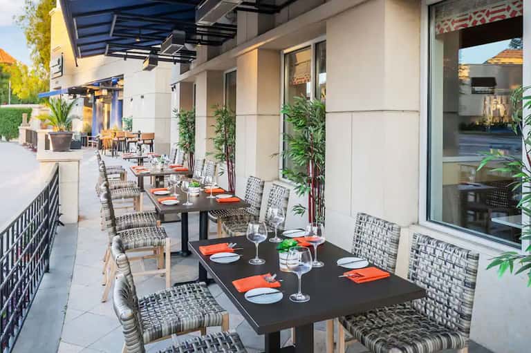 Modern outdoor dining terrace with black tables, patterned chairs, blue shade structures, and plants