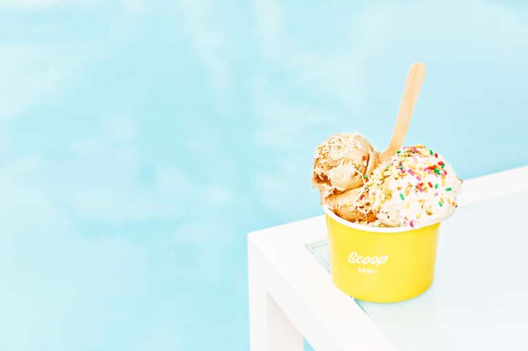 Yellow ice cream cup with two scoops and colorful sprinkles on wooden spoon against bright blue sky