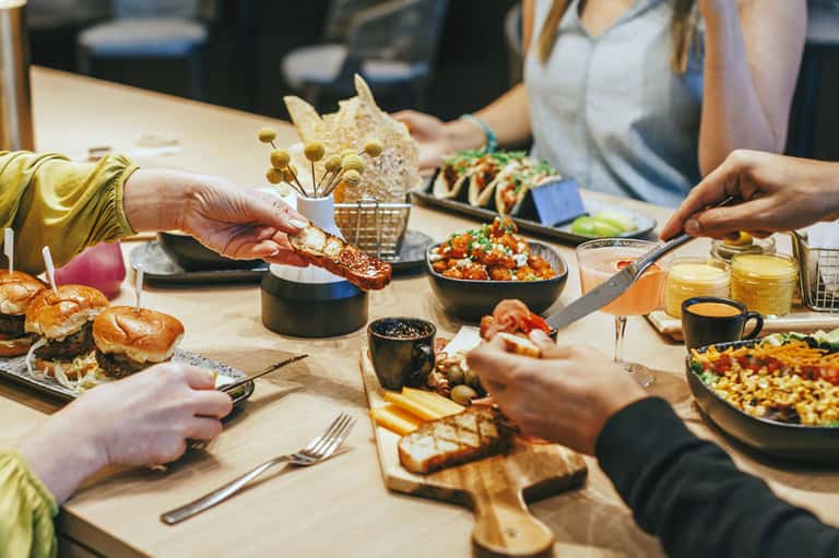 Overhead view of people sharing bread and colorful dishes at a wooden communal dining table