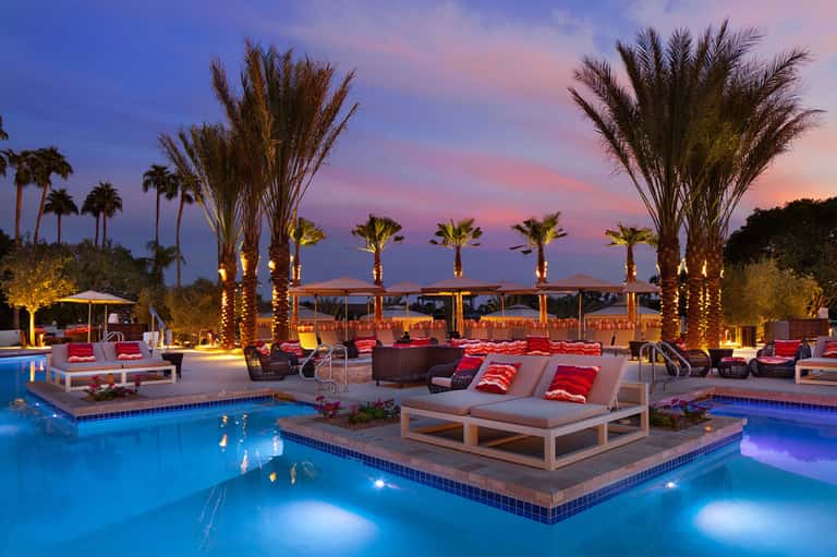 Illuminated resort pool at dusk with loungers, palm trees, and colorful seating area