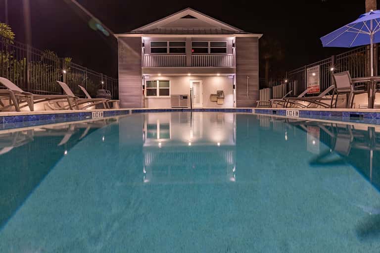 Illuminated resort pool at night with two-story pool house, lounge chairs, blue umbrella, and deck lighting