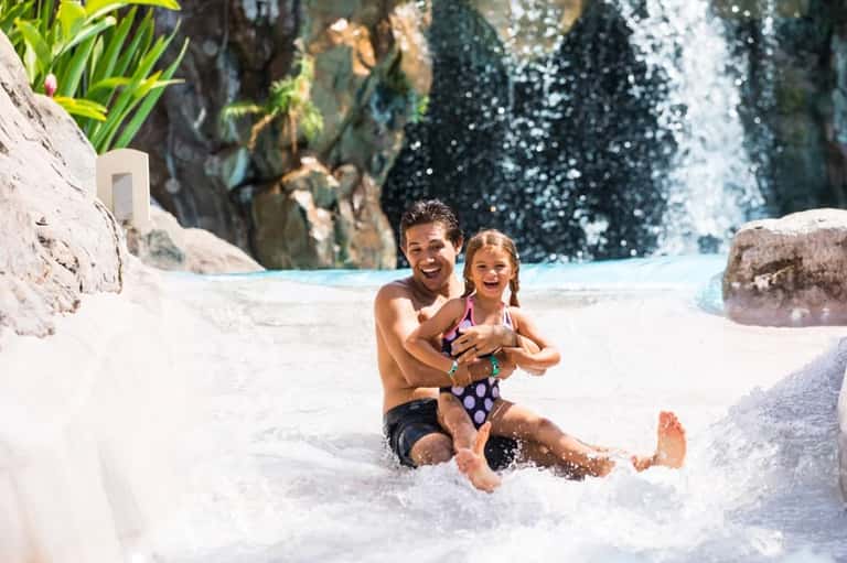 Father and daughter playing in shallow resort pool with waterfall and rock features