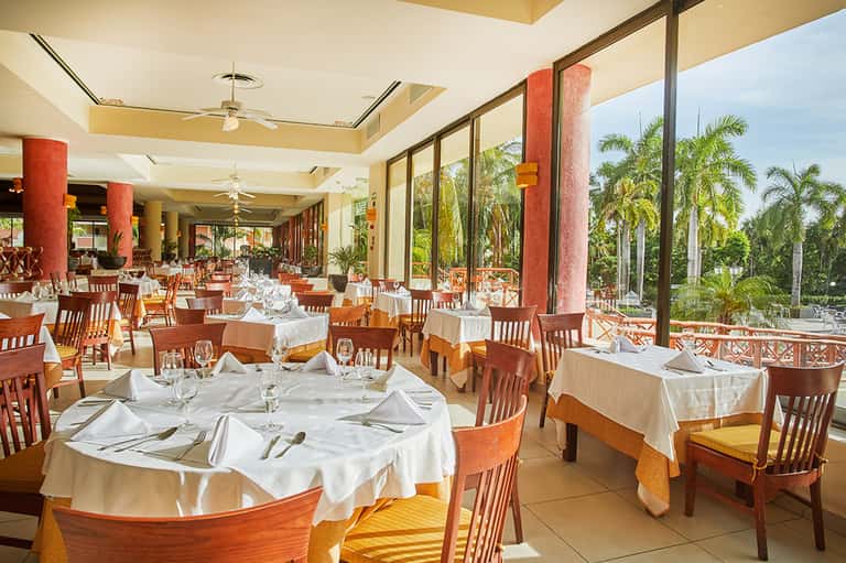 Upscale dining room with white tablecloths, wooden chairs, red columns, and tropical garden views through large windows