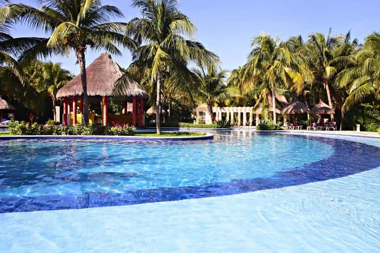 Resort swimming pool with thatched-roof cabanas, palm trees, and tropical landscape