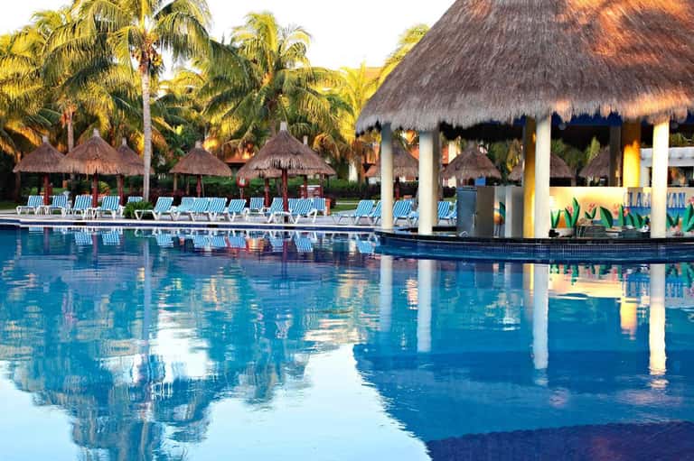 Large resort pool with thatched umbrellas, lounge chairs, palm trees, and pavilion