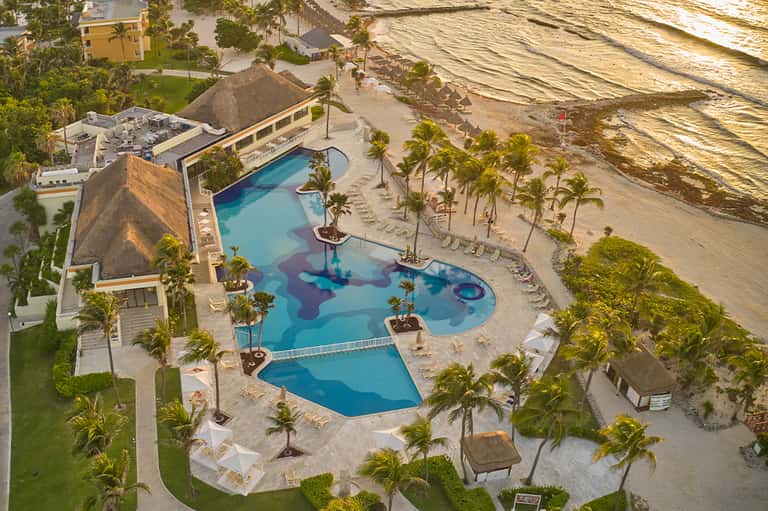 Aerial view of beachfront resort with curved pool, palm trees, and sandy beach landscape