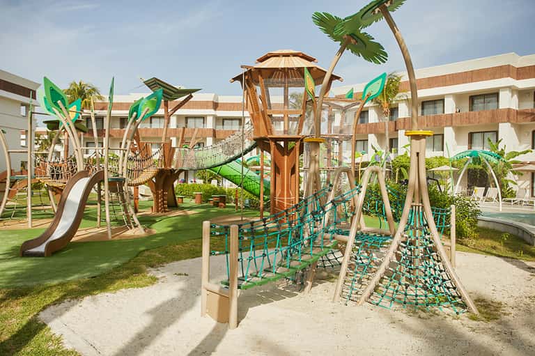 Children's playground with wooden climbing structures, slides, and tropical plant decorations