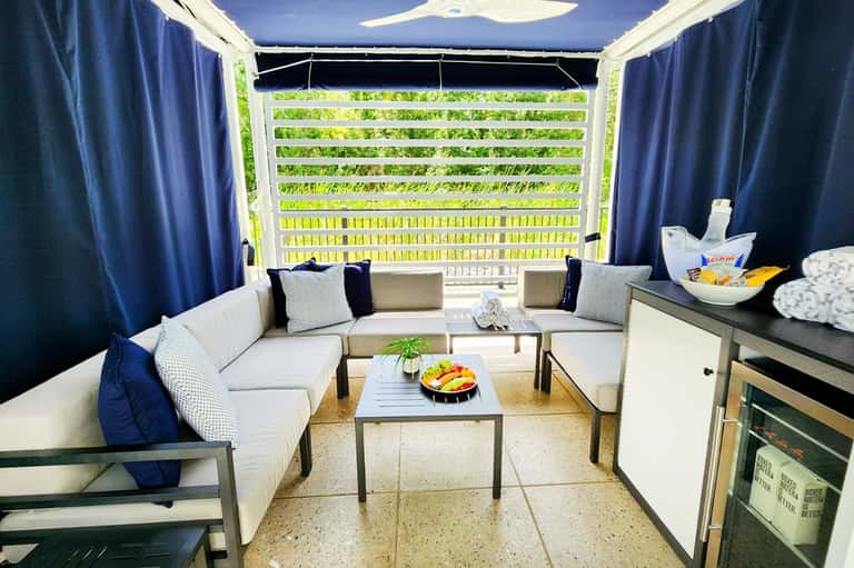 Modern cabana lounge with white sectional sofa, blue curtains, kitchenette, and lush garden view