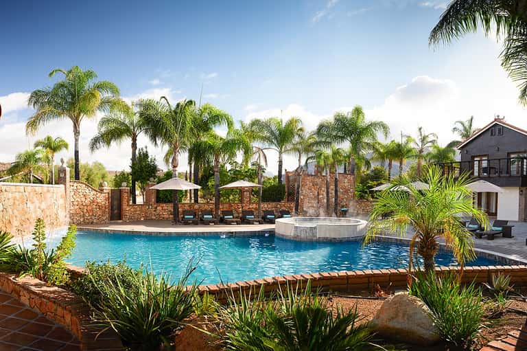 Resort pool with stone wall, palm trees, umbrellas, and Mediterranean-style building with mountain views