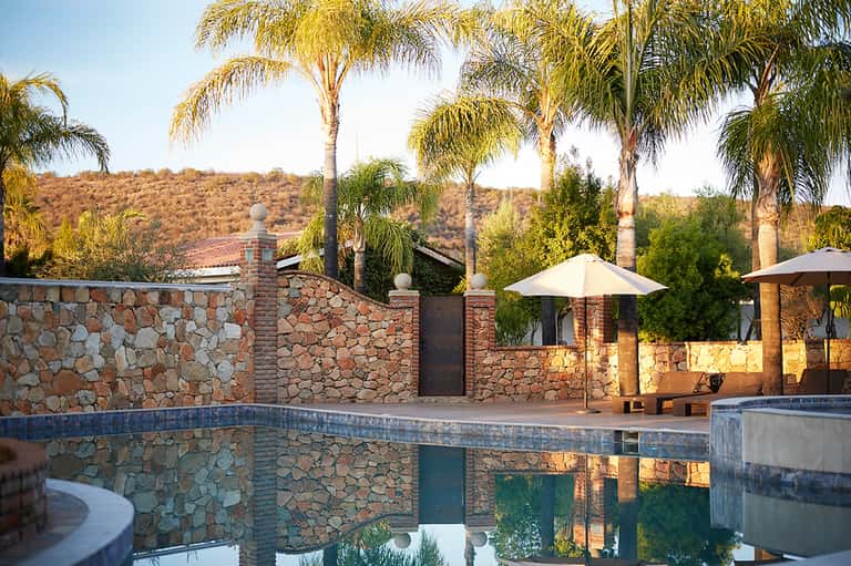Luxury resort pool with stone walls, palm trees, umbrellas, and hillside backdrop
