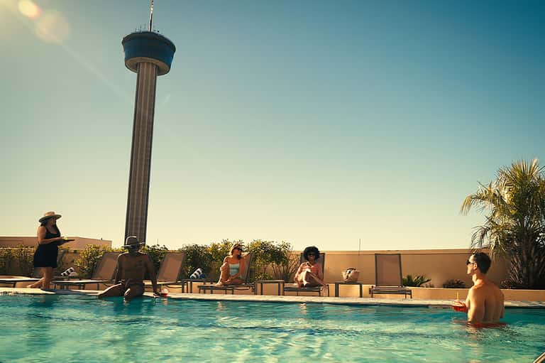 Resort rooftop pool with loungers, guests swimming, observation tower visible, palm trees, and clear blue sky