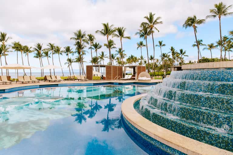 Modern resort pool with waterfall feature, lounge chairs, palm trees, and ocean views