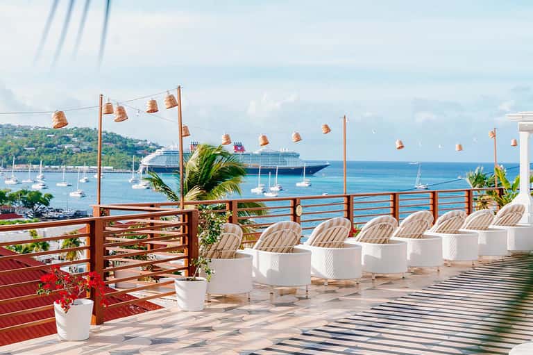Oceanfront terrace with white lounge chairs, bronze pendant lights, wooden railing, and cruise ship in harbor