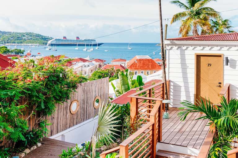 Tropical villa balcony overlooking harbor with cruise ship, colorful buildings, and lush plants