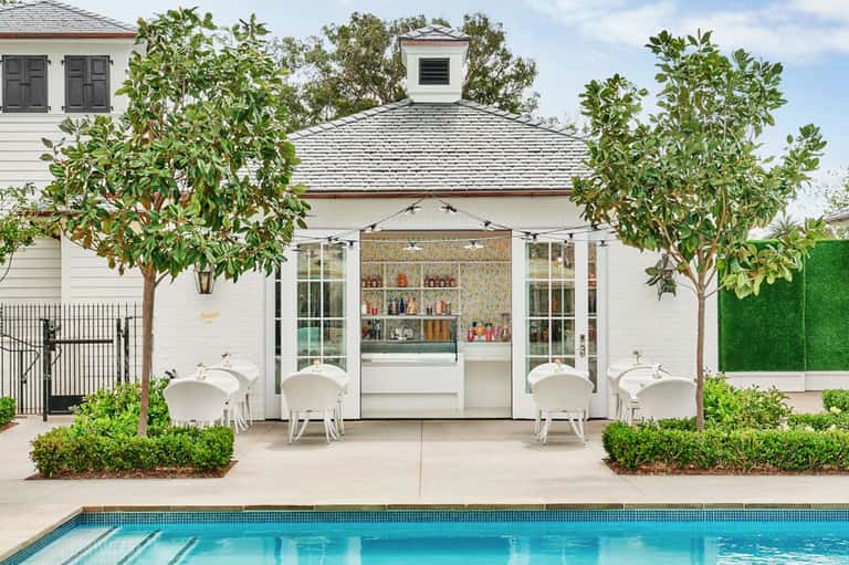 Luxury poolside cabana with white furniture, manicured landscaping, and spa facility featuring glass doors and shelving