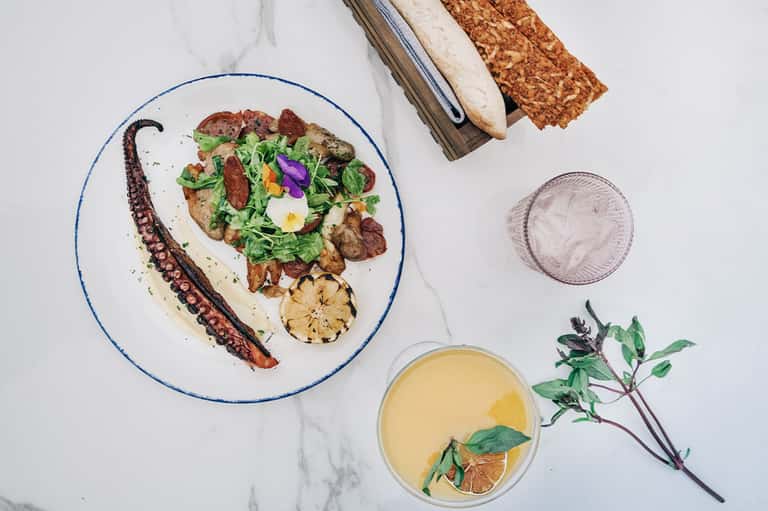 Octopus and salad plated on white marble with bread, juice, and herbs