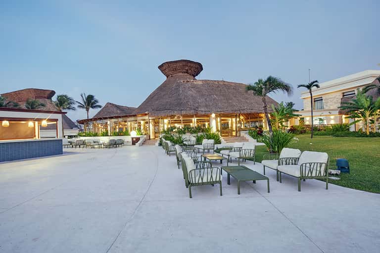 Resort courtyard at dusk with thatched-roof structures, outdoor seating, tropical plants, and modern buildings